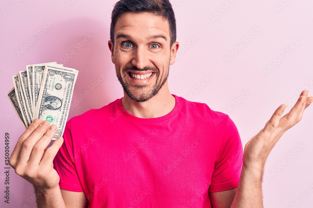 Young handsome man holding bunch of dollars banknotes over isolated pink background celebrating achievement with happy smile and winner expression with raised hand Young handsome man holding bunch of dollars banknotes over isolated pink background celebrating achievement with happy smile and winner expression with raised hand