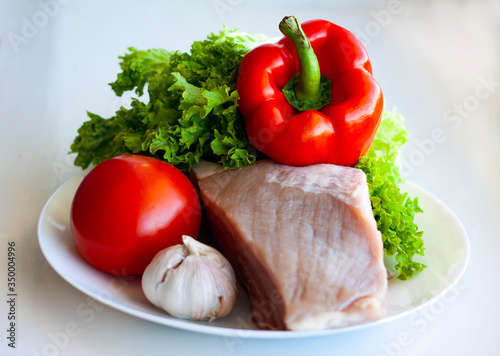 A piece of raw pork on a white plate with lettuce, sweet pepper, tomato and garlic. Healthy eating and cooking at home concept