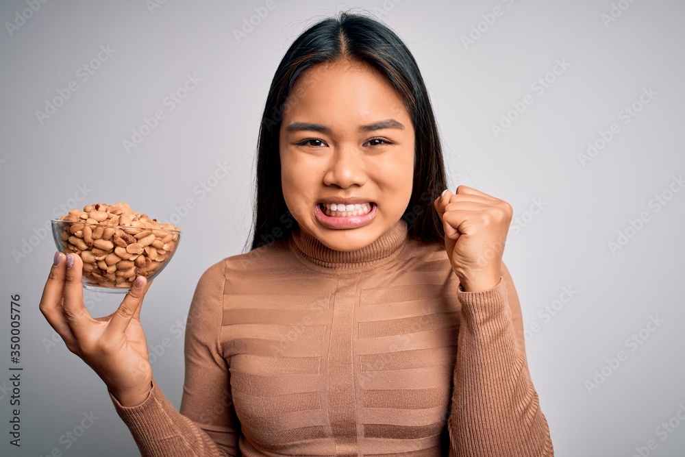 Young asian girl holding bowl of healthy peanuts eating as snack for diet over white background screaming proud and celebrating victory and success very excited, cheering emotion Young asian girl holding bowl of healthy peanuts eating as snack for diet over white background screaming proud and celebrating victory and success very excited, cheering emotion