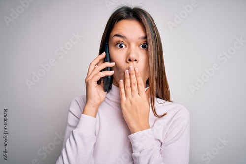 Beautiful brunette girl having conversation talking on the smartphone over white background cover mouth with hand shocked with shame for mistake, expression of fear, scared in silence, secret concept