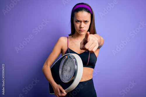 Young beautiful slim sporty girl doing sport wearing sportswear holding weight machine pointing with finger to the camera and to you  hand sign  positive and confident gesture from the front