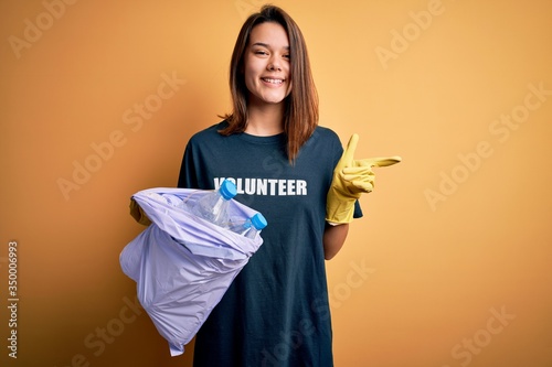 Beautiful volunteer girl caring environment doing volunteering holding bag with rubish bottles very happy pointing with hand and finger to the side