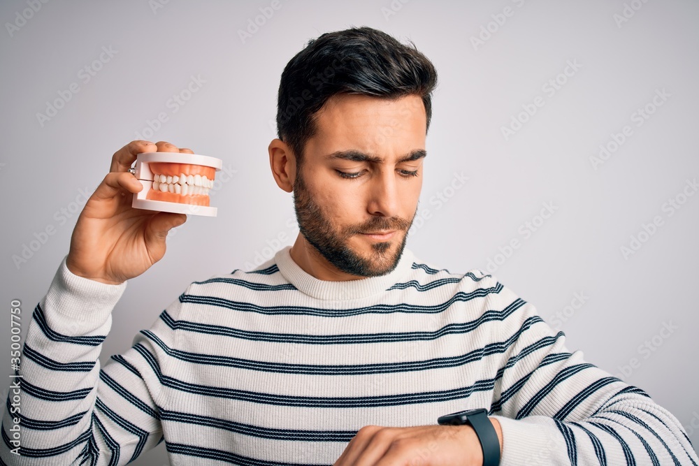 Young handsome man with beard holding plastic denture teeth over white background Checking the time on wrist watch, relaxed and confident Young handsome man with beard holding plastic denture teeth over white background Checking the time on wrist watch, relaxed and confident