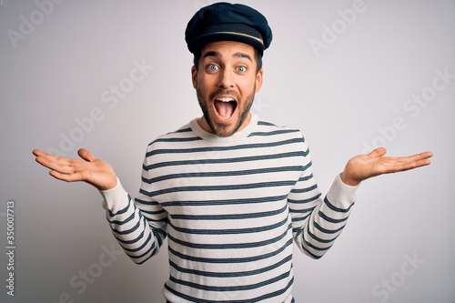 Young handsome sailor man with beard wearing navy striped uniform and captain hat celebrating crazy and amazed for success with arms raised and open eyes screaming excited. Winner concept