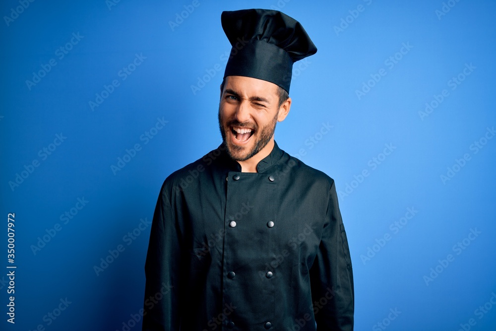 Young handsome chef man with beard wearing cooker uniform and hat over blue background winking looking at the camera with sexy expression, cheerful and happy face. Young handsome chef man with beard wearing cooker uniform and hat over blue background winking looking at the camera with sexy expression, cheerful and happy face.