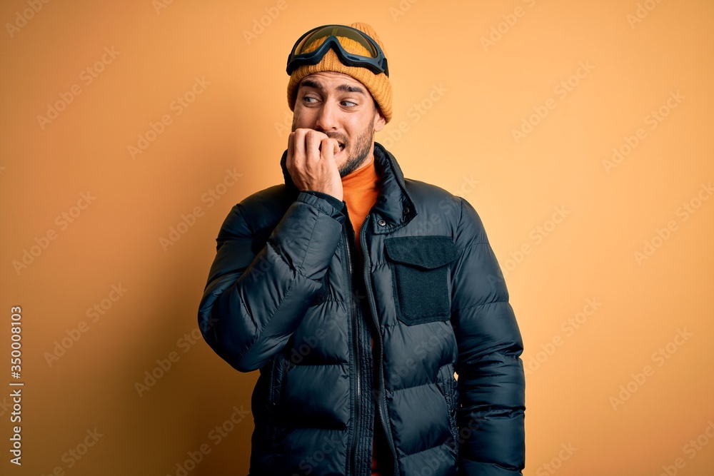 Young handsome skier man with beard wearing snow sportswear and ski goggles looking stressed and nervous with hands on mouth biting nails. Anxiety problem. Young handsome skier man with beard wearing snow sportswear and ski goggles looking stressed and nervous with hands on mouth biting nails. Anxiety problem.