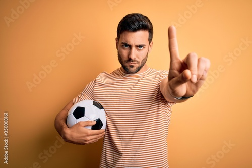 Handsome player man with beard playing soccer holding footballl ball over yellow background Pointing with finger up and angry expression, showing no gesture