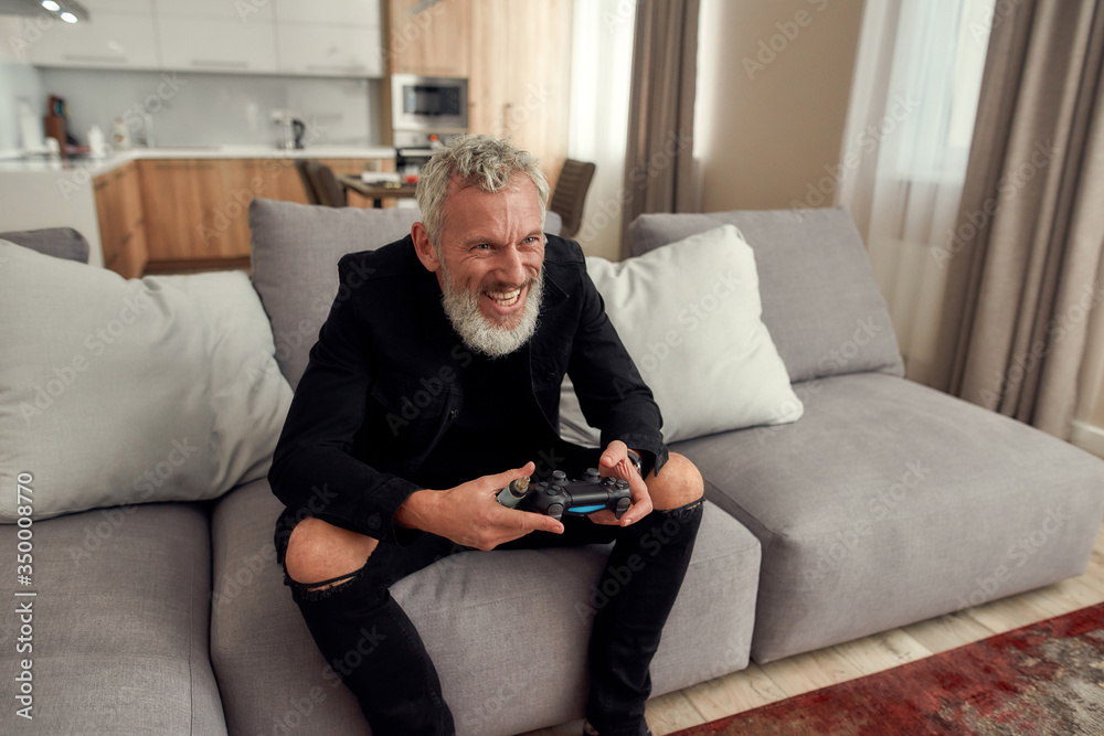 Bringing Game to Life. Happy bearded middle-aged man holding weed vaporizer, dry herb vape pen while playing video games, sitting on the couch at home
