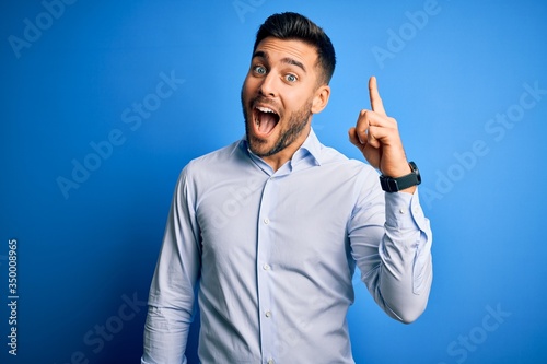 Young handsome man wearing elegant shirt standing over isolated blue background pointing finger up with successful idea. Exited and happy. Number one.
