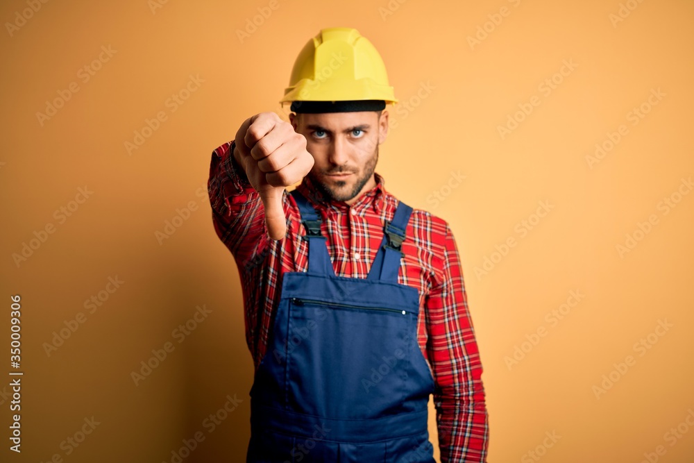 Young builder man wearing construction uniform and safety helmet over yellow isolated background looking unhappy and angry showing rejection and negative with thumbs down gesture. Bad expression. Young builder man wearing construction uniform and safety helmet over yellow isolated background looking unhappy and angry showing rejection and negative with thumbs down gesture. Bad expression.