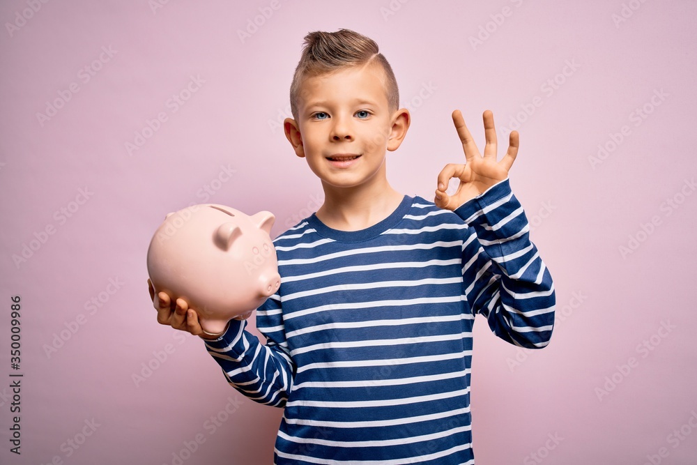 Young little caucasian kid banking money on piggy bank as wealth savings over pink background doing ok sign with fingers, excellent symbol