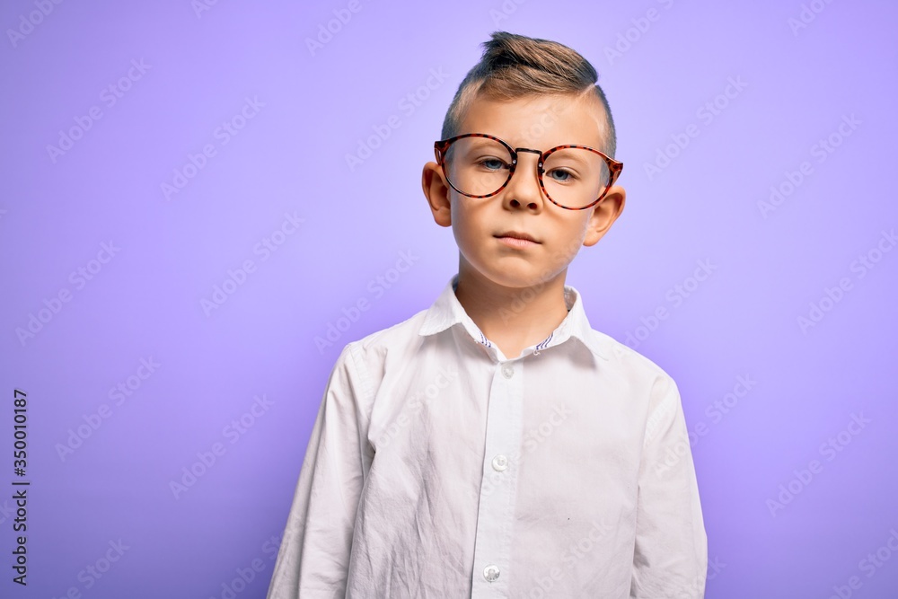 Young little caucasian kid with blue eyes wearing glasses and white shirt over purple background with serious expression on face. Simple and natural looking at the camera.