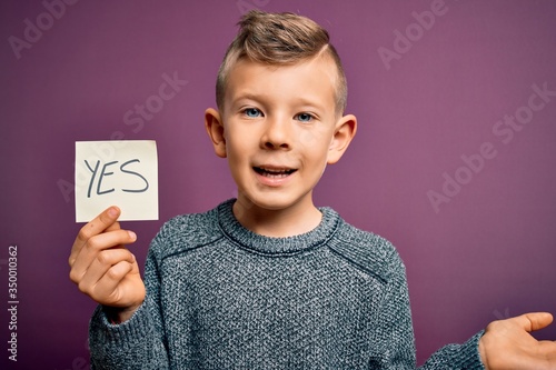 Young little caucasian kid showing YES word on a paper note as positive and success message very happy and excited, winner expression celebrating victory screaming with big smile and raised hands