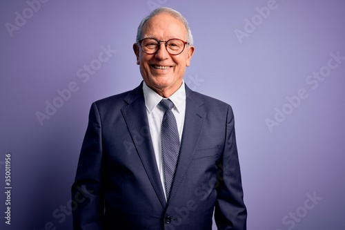 Grey haired senior business man wearing glasses and elegant suit and tie over purple background with a happy and cool smile on face. Lucky person.
