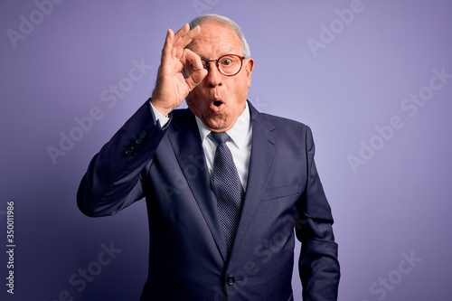 Grey haired senior business man wearing glasses and elegant suit and tie over purple background doing ok gesture shocked with surprised face, eye looking through fingers. Unbelieving expression.