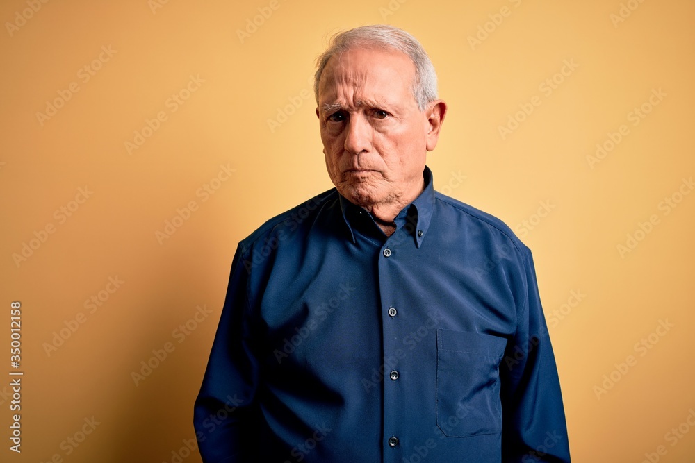 Grey haired senior man wearing casual blue shirt standing over yellow background skeptic and nervous, frowning upset because of problem. Negative person. Grey haired senior man wearing casual blue shirt standing over yellow background skeptic and nervous, frowning upset because of problem. Negative person.