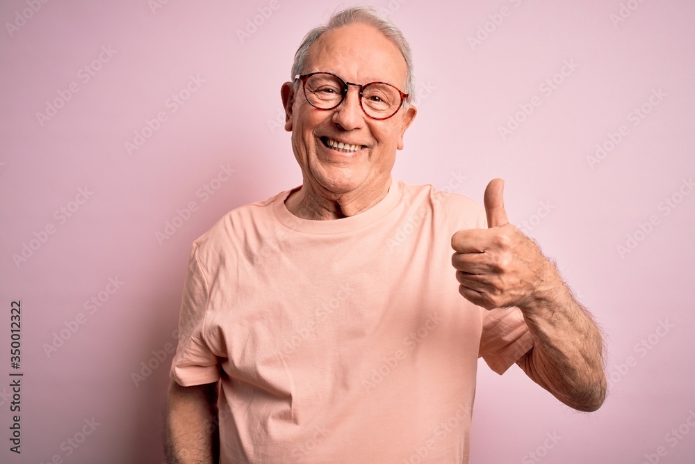 Grey haired senior man wearing glasses standing over pink isolated background doing happy thumbs up gesture with hand. Approving expression looking at the camera showing success. Grey haired senior man wearing glasses standing over pink isolated background doing happy thumbs up gesture with hand. Approving expression looking at the camera showing success.