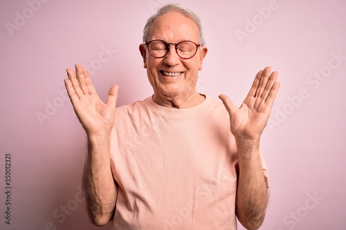 Grey haired senior man wearing glasses standing over pink isolated background celebrating mad and crazy for success with arms raised and closed eyes screaming excited. Winner concept