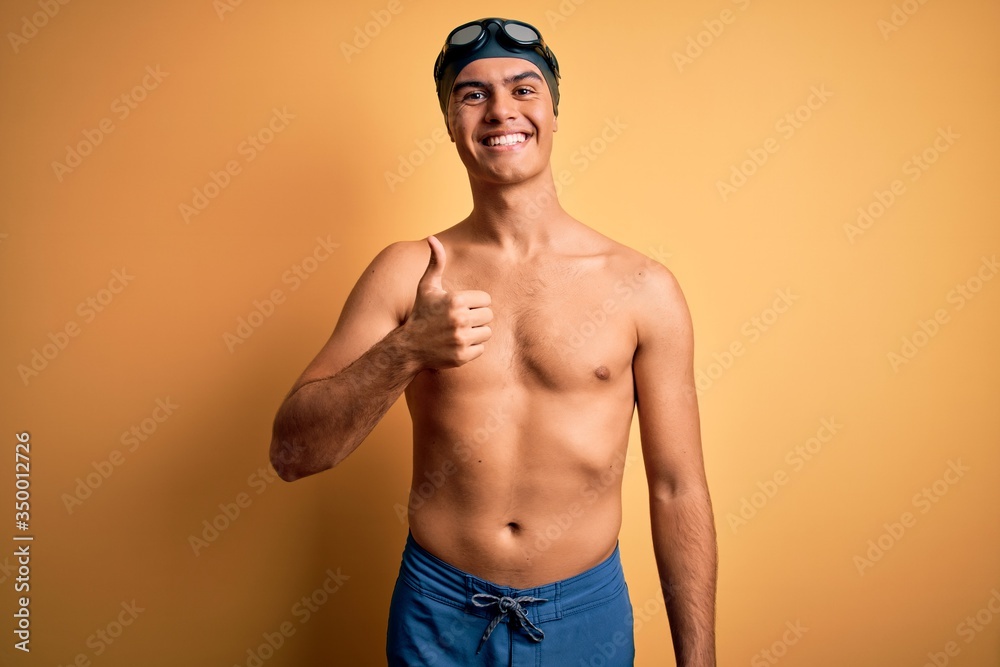 Young handsome man shirtless wearing swimsuit and swim cap over isolated yellow background doing happy thumbs up gesture with hand. Approving expression looking at the camera showing success.