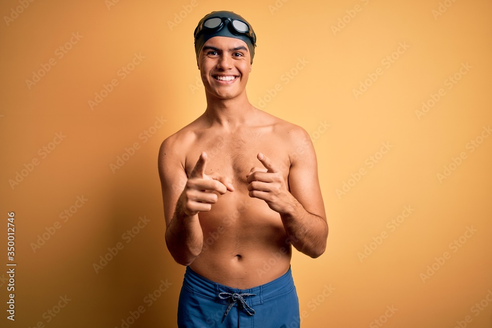 Young handsome man shirtless wearing swimsuit and swim cap over isolated yellow background pointing fingers to camera with happy and funny face. Good energy and vibes.