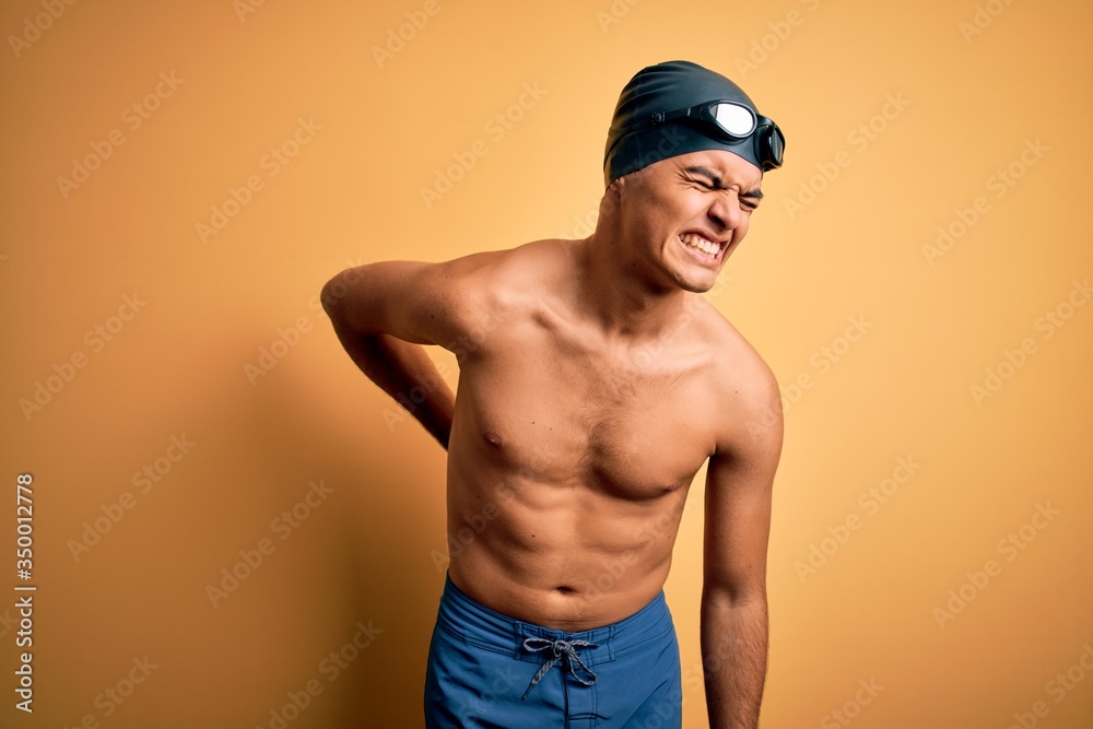 Young handsome man shirtless wearing swimsuit and swim cap over isolated yellow background Suffering of backache, touching back with hand, muscular pain