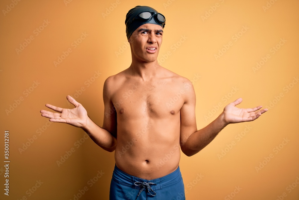 Young handsome man shirtless wearing swimsuit and swim cap over isolated yellow background clueless and confused with open arms, no idea concept.