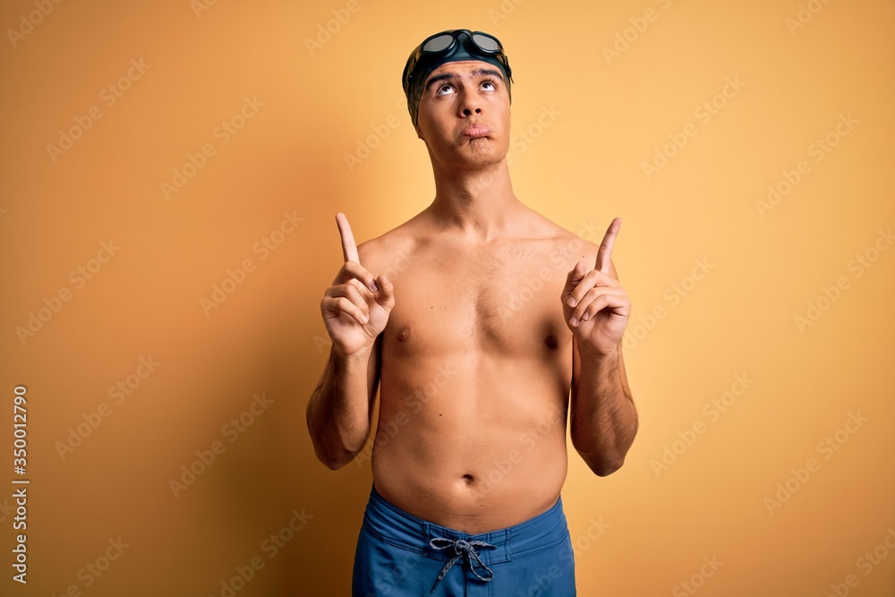 Young handsome man shirtless wearing swimsuit and swim cap over isolated yellow background Pointing up looking sad and upset, indicating direction with fingers, unhappy and depressed.