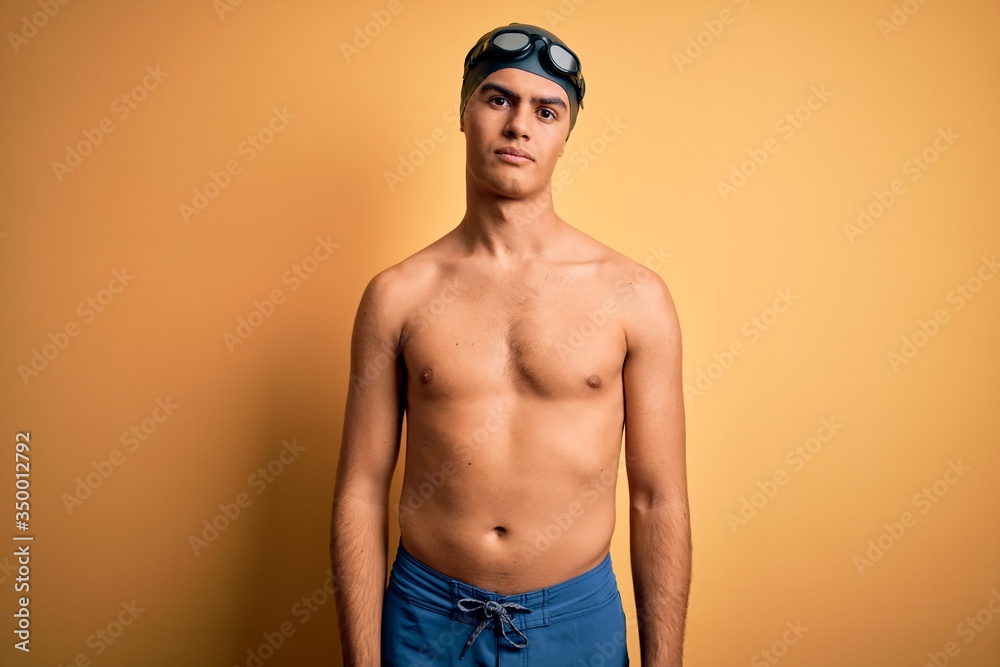 Young handsome man shirtless wearing swimsuit and swim cap over isolated yellow background Relaxed with serious expression on face. Simple and natural looking at the camera.