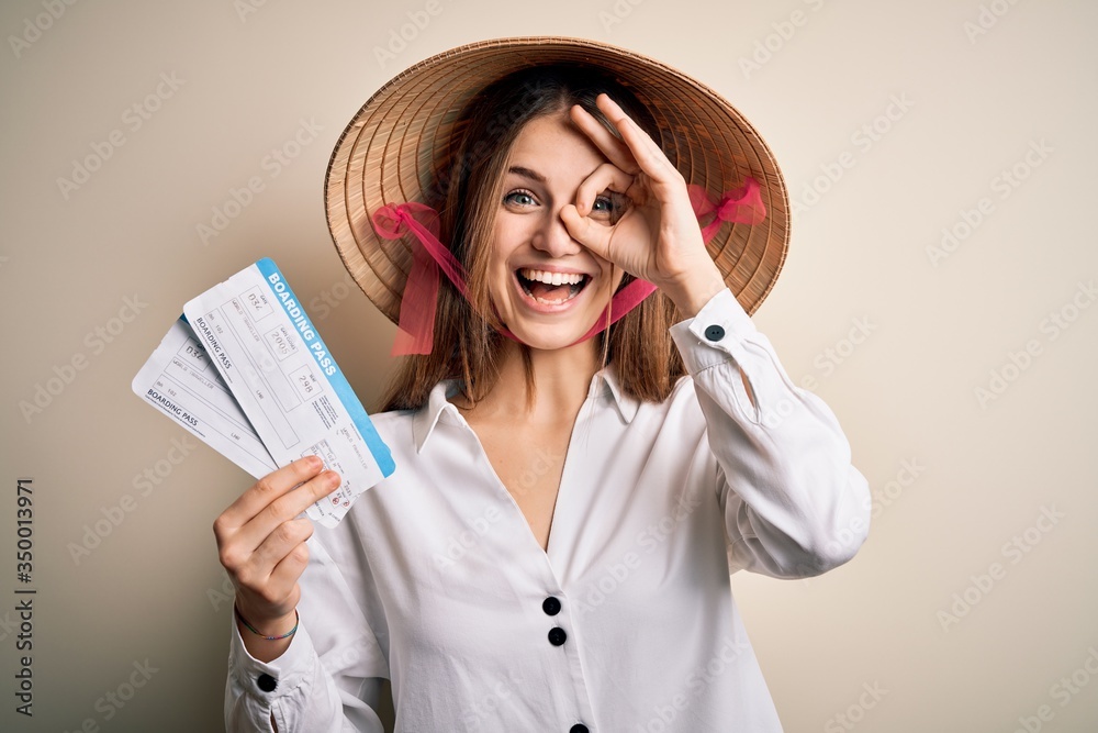 Young beautiful redhead tourist woman wearing asian traditional hat holding boarding pass with happy face smiling doing ok sign with hand on eye looking through fingers Young beautiful redhead tourist woman wearing asian traditional hat holding boarding pass with happy face smiling doing ok sign with hand on eye looking through fingers