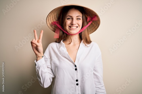 Young beautiful redhead woman wearing asian traditional conical hat over white background smiling with happy face winking at the camera doing victory sign. Number two.