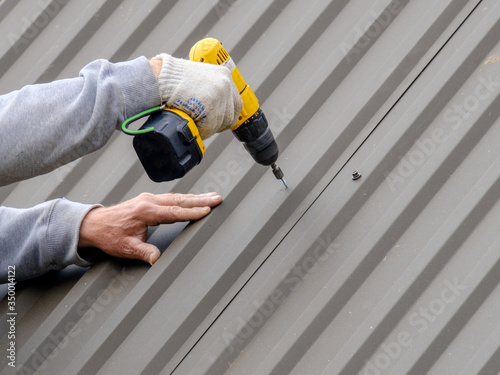 Men's hands in work gloves with a yellow screwdriver screw the roofing sheet to the roof of a country house. Cordless drill. The use of electrical engineering and technology.