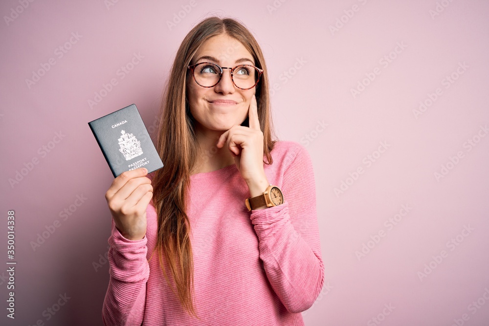 Beautiful redhead tourist woman holding canadian canada passport over pink bakcground serious face thinking about question, very confused idea