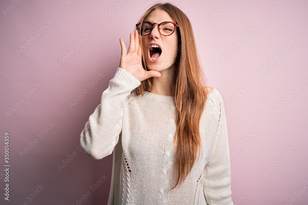 Young beautiful redhead woman wearing casual sweater and glasses over pink background shouting and screaming loud to side with hand on mouth. Communication concept.