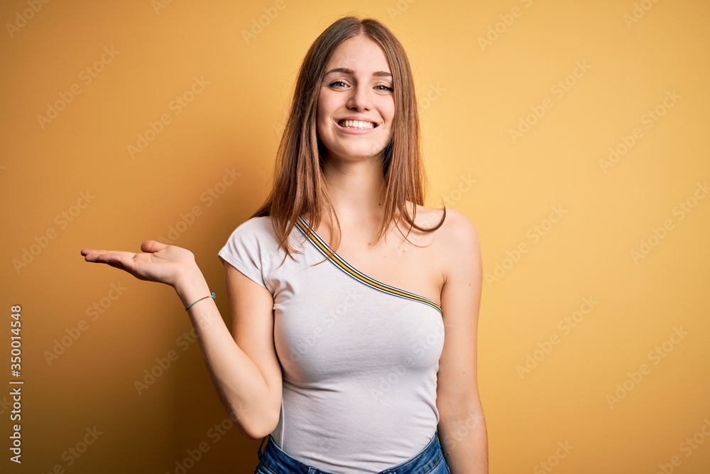 Young beautiful redhead woman wearing casual t-shirt over isolated yellow background smiling cheerful presenting and pointing with palm of hand looking at the camera. Young beautiful redhead woman wearing casual t-shirt over isolated yellow background smiling cheerful presenting and pointing with palm of hand looking at the camera.