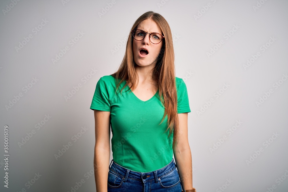Obraz premium Young beautiful redhead woman wearing casual green t-shirt and glasses over white background In shock face, looking skeptical and sarcastic, surprised with open mouth