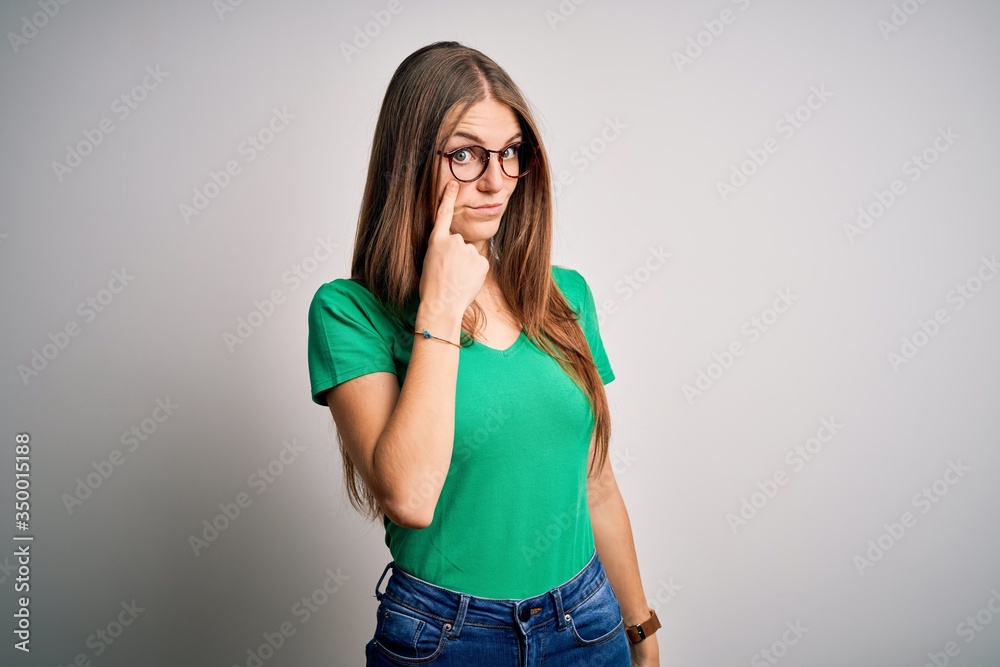 Young beautiful redhead woman wearing casual green t-shirt and glasses over white background Pointing to the eye watching you gesture, suspicious expression Young beautiful redhead woman wearing casual green t-shirt and glasses over white background Pointing to the eye watching you gesture, suspicious expression