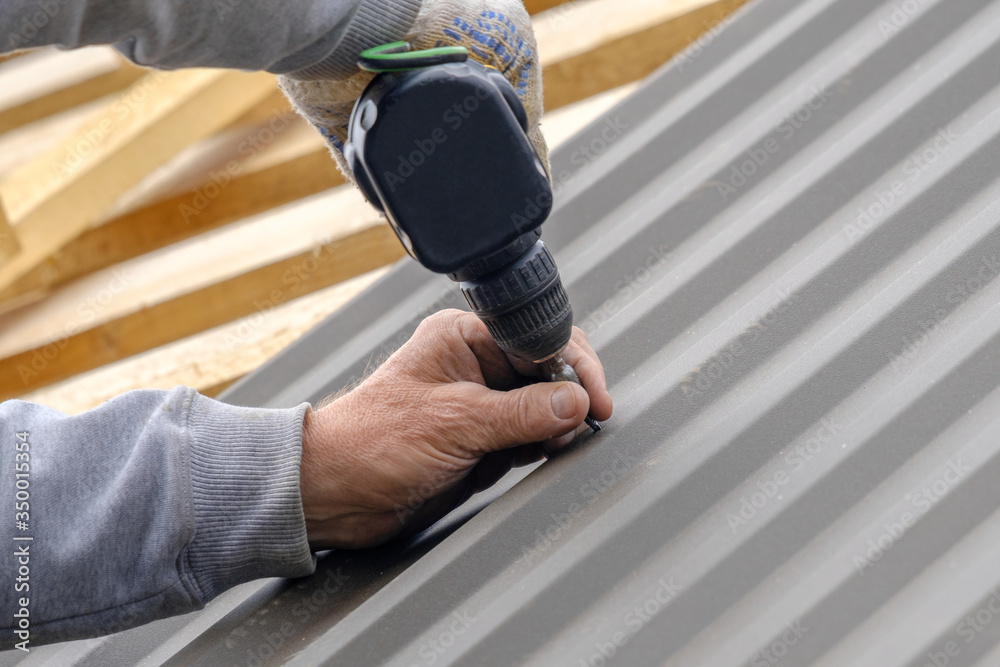 Men's hands in work gloves with a yellow screwdriver screw the roofing sheet to the roof of a country house. Cordless drill. The use of electrical engineering and technology.