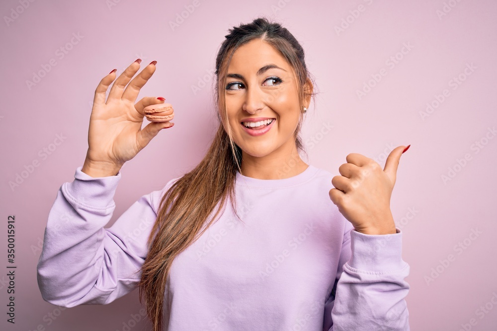 Young beautiful woman eating french pastry sweet macaron over pink isolated background pointing and showing with thumb up to the side with happy face smiling Young beautiful woman eating french pastry sweet macaron over pink isolated background pointing and showing with thumb up to the side with happy face smiling