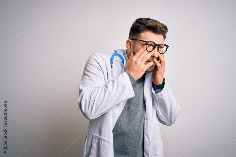 Young doctor man with blue eyes wearing medical coat and stethoscope over isolated background rubbing eyes for fatigue and headache, sleepy and tired expression. Vision problem Young doctor man with blue eyes wearing medical coat and stethoscope over isolated background rubbing eyes for fatigue and headache, sleepy and tired expression. Vision problem