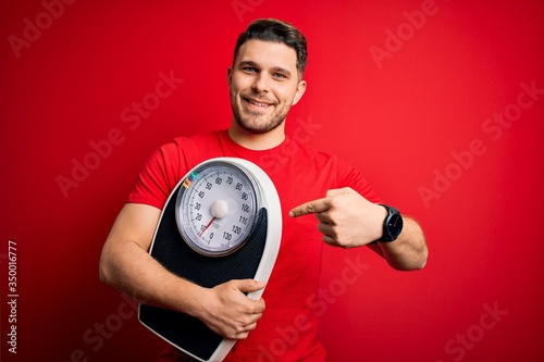 Young fitness man with blue eyes holding scale dieting for healthy weight over red background very happy pointing with hand and finger
