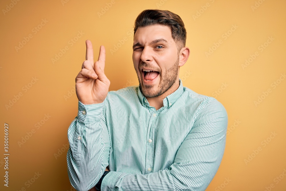 Young business man with blue eyes wearing elegant green shirt over yellow background smiling with happy face winking at the camera doing victory sign. Number two. Young business man with blue eyes wearing elegant green shirt over yellow background smiling with happy face winking at the camera doing victory sign. Number two.