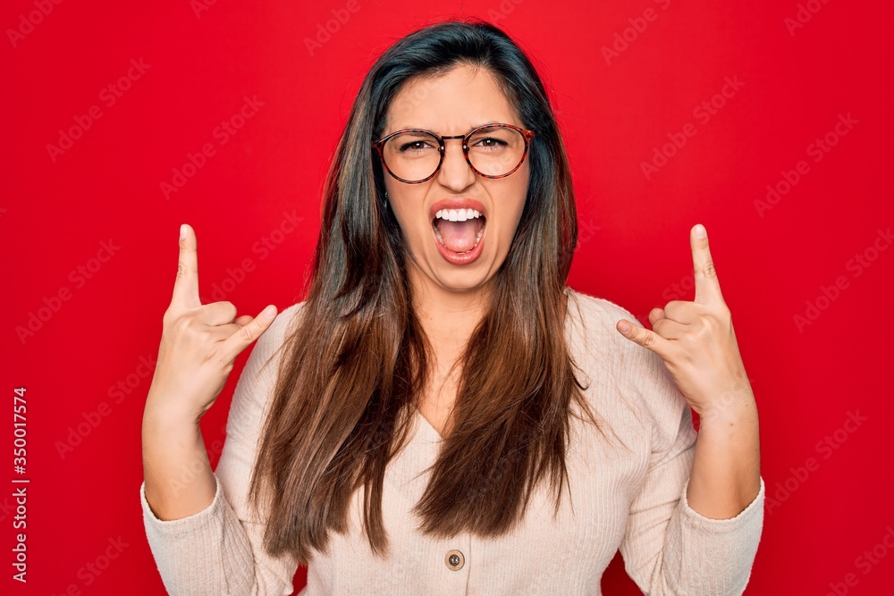 Young hispanic smart woman wearing glasses standing over red isolated background shouting with crazy expression doing rock symbol with hands up. Music star. Heavy concept. Young hispanic smart woman wearing glasses standing over red isolated background shouting with crazy expression doing rock symbol with hands up. Music star. Heavy concept.