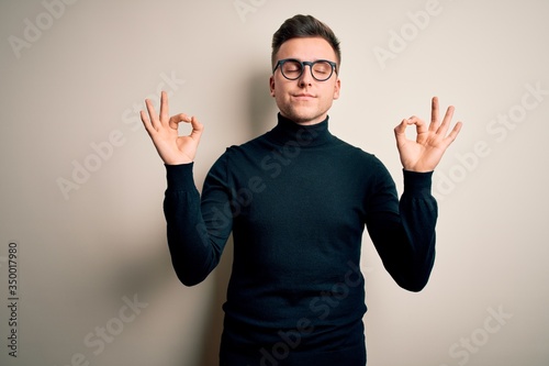 Young handsome caucasian man wearing glasses and casual sweater over isolated background relax and smiling with eyes closed doing meditation gesture with fingers. Yoga concept. © Krakenimages.com