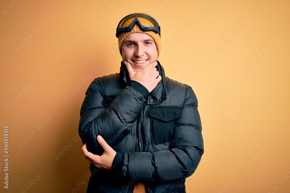Young handsome caucasian man wearing hat, coat and ski glasses for winter and snow weather looking confident at the camera smiling with crossed arms and hand raised on chin. Thinking positive. Young handsome caucasian man wearing hat, coat and ski glasses for winter and snow weather looking confident at the camera smiling with crossed arms and hand raised on chin. Thinking positive.