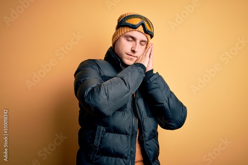 Young handsome caucasian man wearing hat, coat and ski glasses for winter and snow weather sleeping tired dreaming and posing with hands together while smiling with closed eyes. © Krakenimages.com