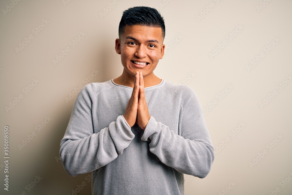 Young handsome latin man wearing casual sweater standing over isolated white background praying with hands together asking for forgiveness smiling confident. Young handsome latin man wearing casual sweater standing over isolated white background praying with hands together asking for forgiveness smiling confident.