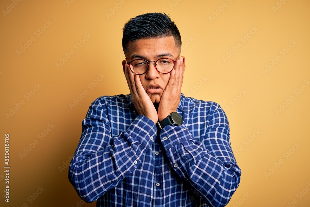 Obraz premium Young handsome latin man wearing casual shirt and glasses over yellow background Tired hands covering face, depression and sadness, upset and irritated for problem