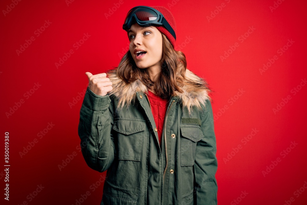 Young blonde girl wearing ski glasses and winter coat for ski weather over red background smiling with happy face looking and pointing to the side with thumb up.