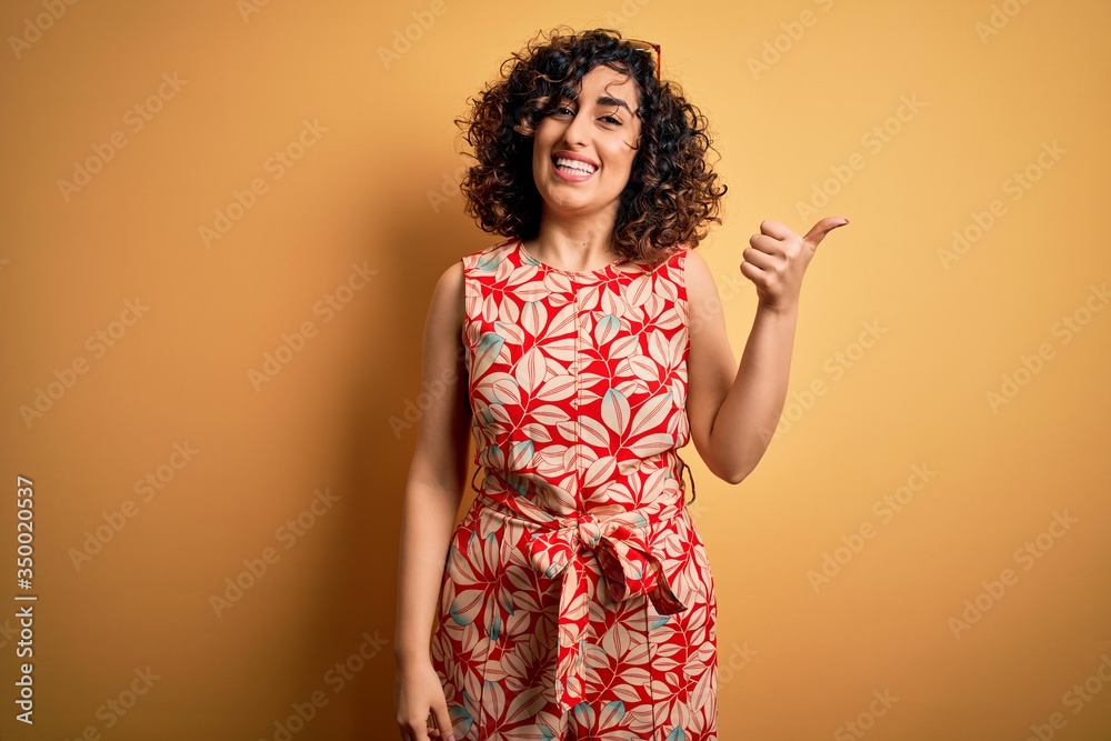 Young beautiful curly arab woman on vacation wearing summer floral dress and sunglasses smiling with happy face looking and pointing to the side with thumb up. Young beautiful curly arab woman on vacation wearing summer floral dress and sunglasses smiling with happy face looking and pointing to the side with thumb up.