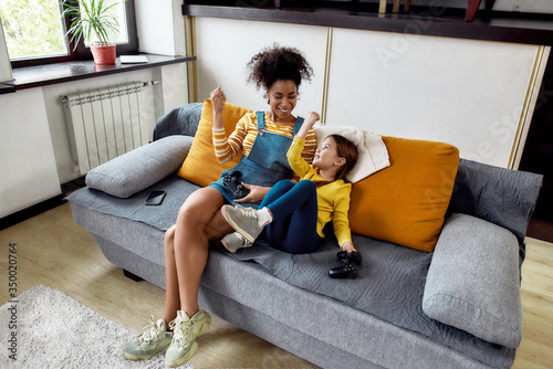 Winners. African american woman, baby sitter and caucasian cute little girl having fun together, playing video games, sitting on the couch. Children education, leisure activities, babysitting concept